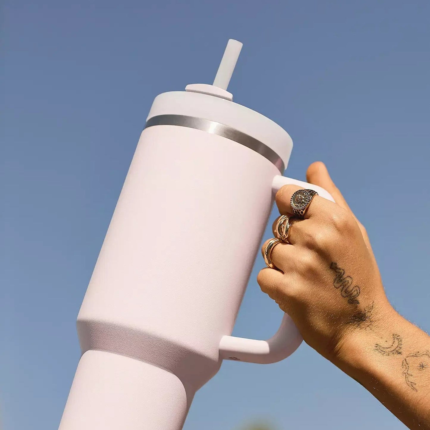 A hand holding a large pink insulated tumbler with a handle and a straw, against a blue sky.